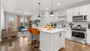 A modern kitchen with white cabinetry, a central island, stainless steel appliances, and a dining area with a blue chair. - The Garden Luxe Retreat (Winter Garden)