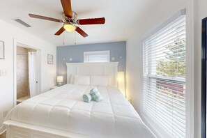 Une chambre à coucher comprenant un lit, un ventilateur de plafond, une fenêtre avec des stores et une porte donnant sur une salle de bain.