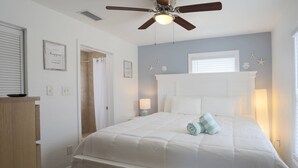 Een slaapkamer met een wit bed, een plafondventilator, een raam met zonwering en een wandklok.