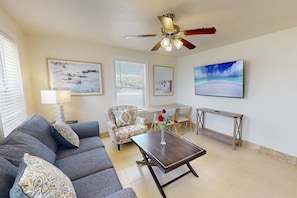 A living room with a sofa, armchair, coffee table, and a ceiling fan. There are framed pictures on the walls and a TV on the wall.