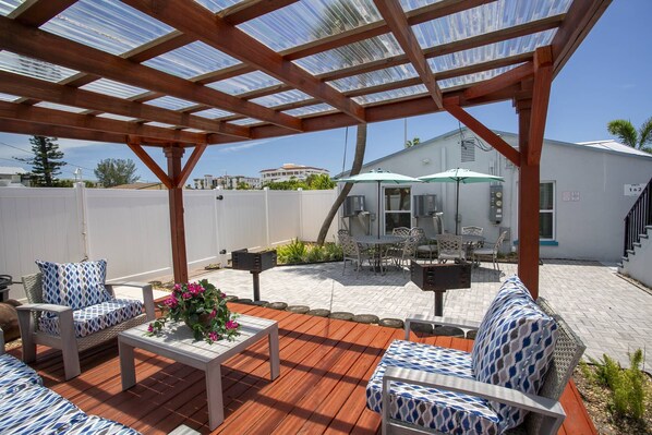 A covered patio area with outdoor furniture, a table with a flower centerpiece, and a white building in the background.