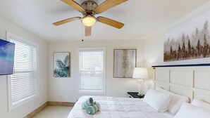 A bedroom with a bed, a ceiling fan, a nightstand, a lamp, and artwork on the walls.