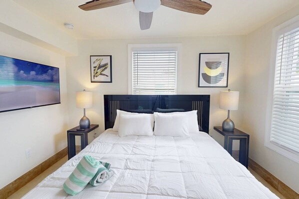 A bedroom with a large bed, two bedside tables with lamps, a ceiling fan, and artwork on the walls.