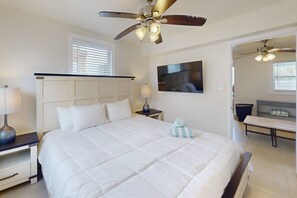 Ein Schlafzimmer mit einem Bett, einem Deckenventilator, einem Nachttisch mit Lampe, einem an der Wand montierten Fernseher und einem durch eine geöffnete Tür einsehbaren Badezimmer.