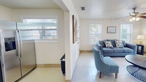 A modern kitchen with a built-in refrigerator and oven, a grey sofa with patterned cushions, and a dining area with a round table and a ceiling fan.