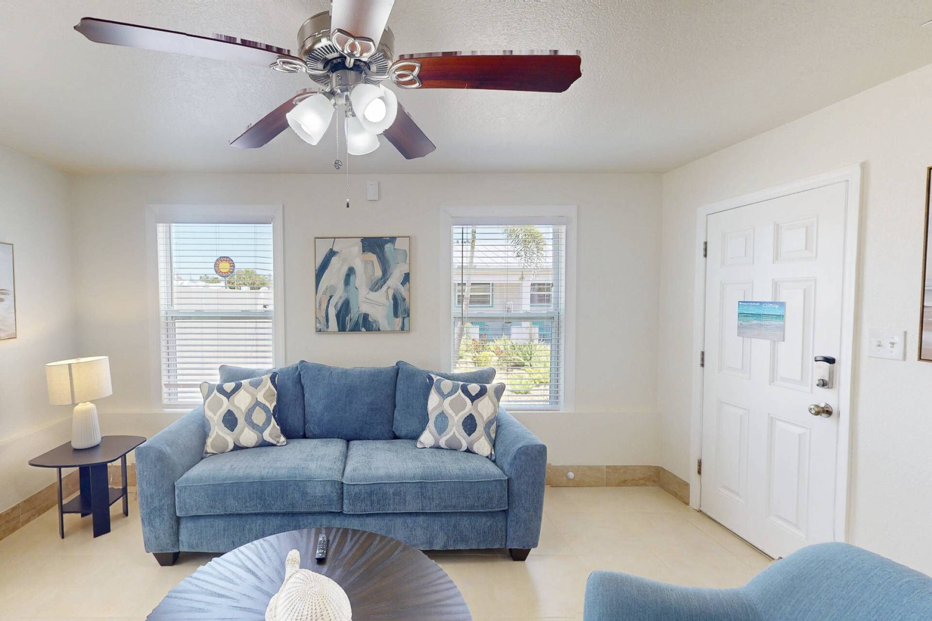 A living room with a blue sofa, a coffee table, a ceiling fan, and two windows with blinds.