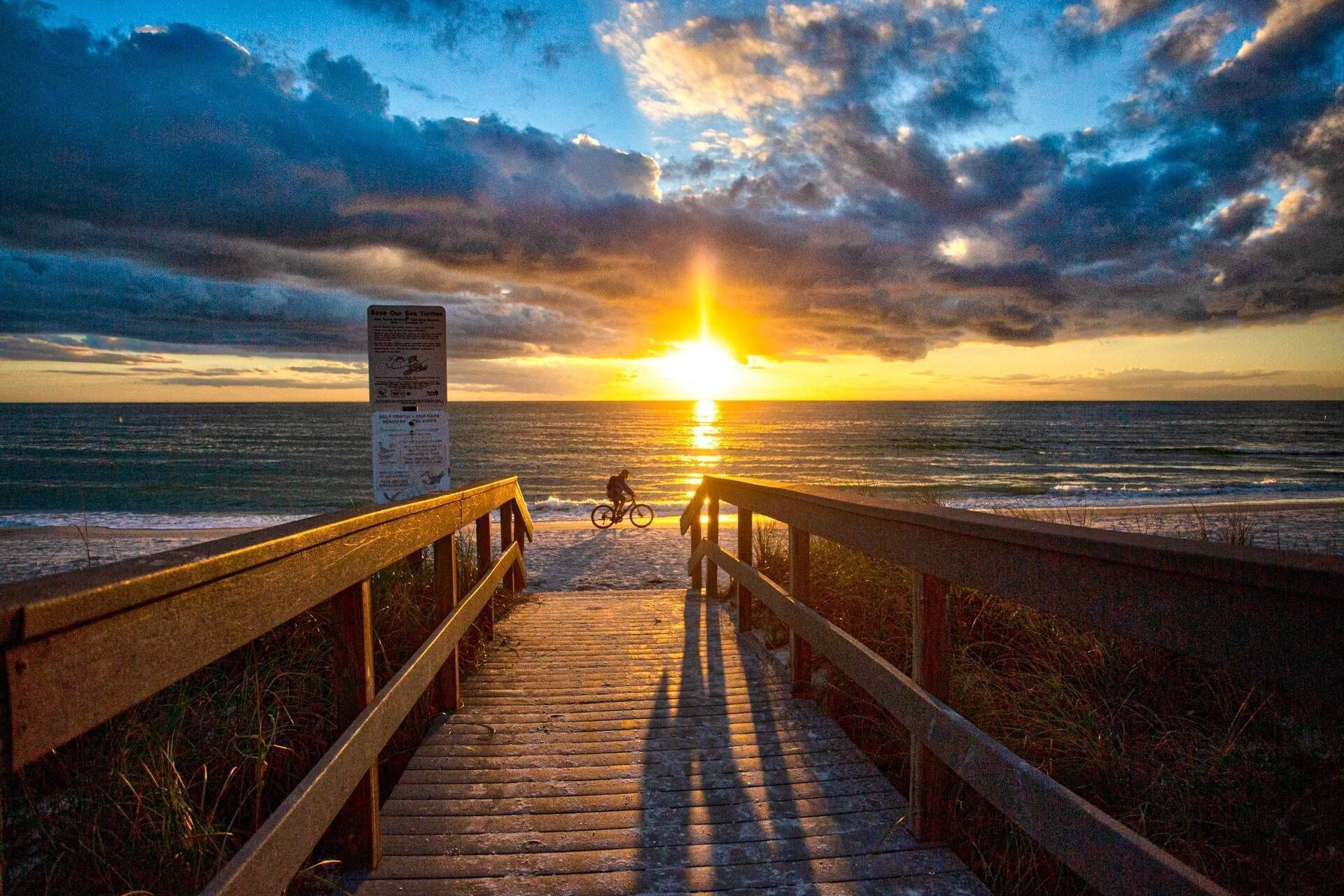 Ein Holzsteg führt zum Meer, im Hintergrund ist ein Sonnenuntergang zu sehen.