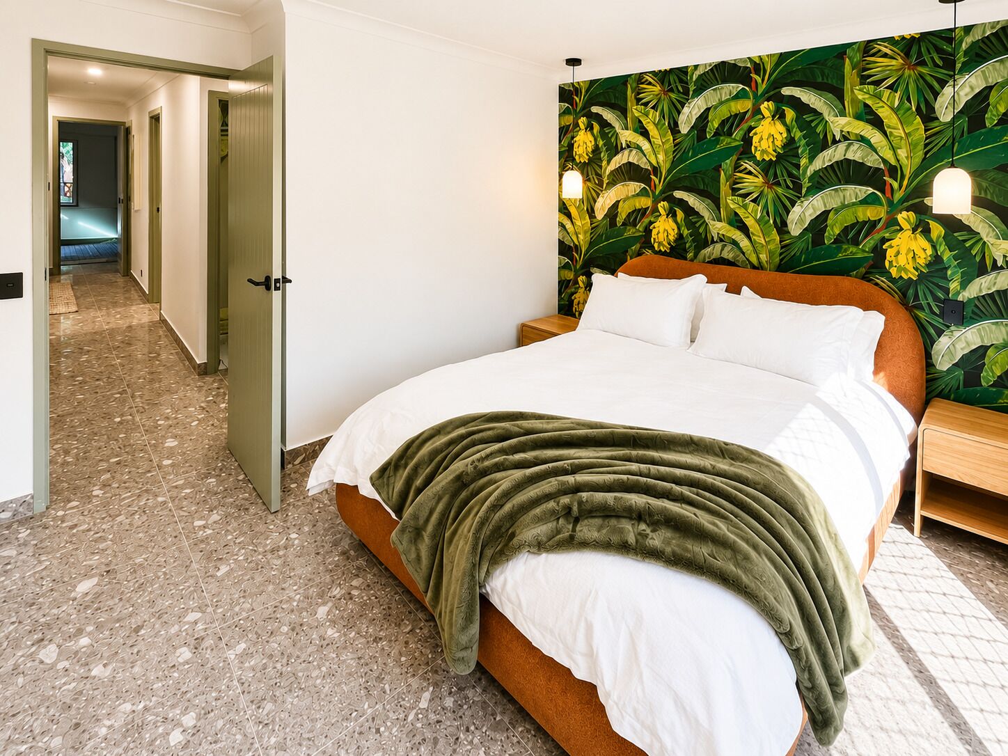 Une chambre à coucher avec un lit, une couverture verte, une table de chevet et un mur orné d’un motif de feuilles tropicales.