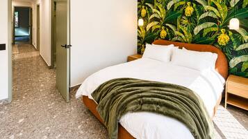 Une chambre à coucher avec un lit, une couverture verte, une table de chevet et un mur orné d’un motif de feuilles tropicales.