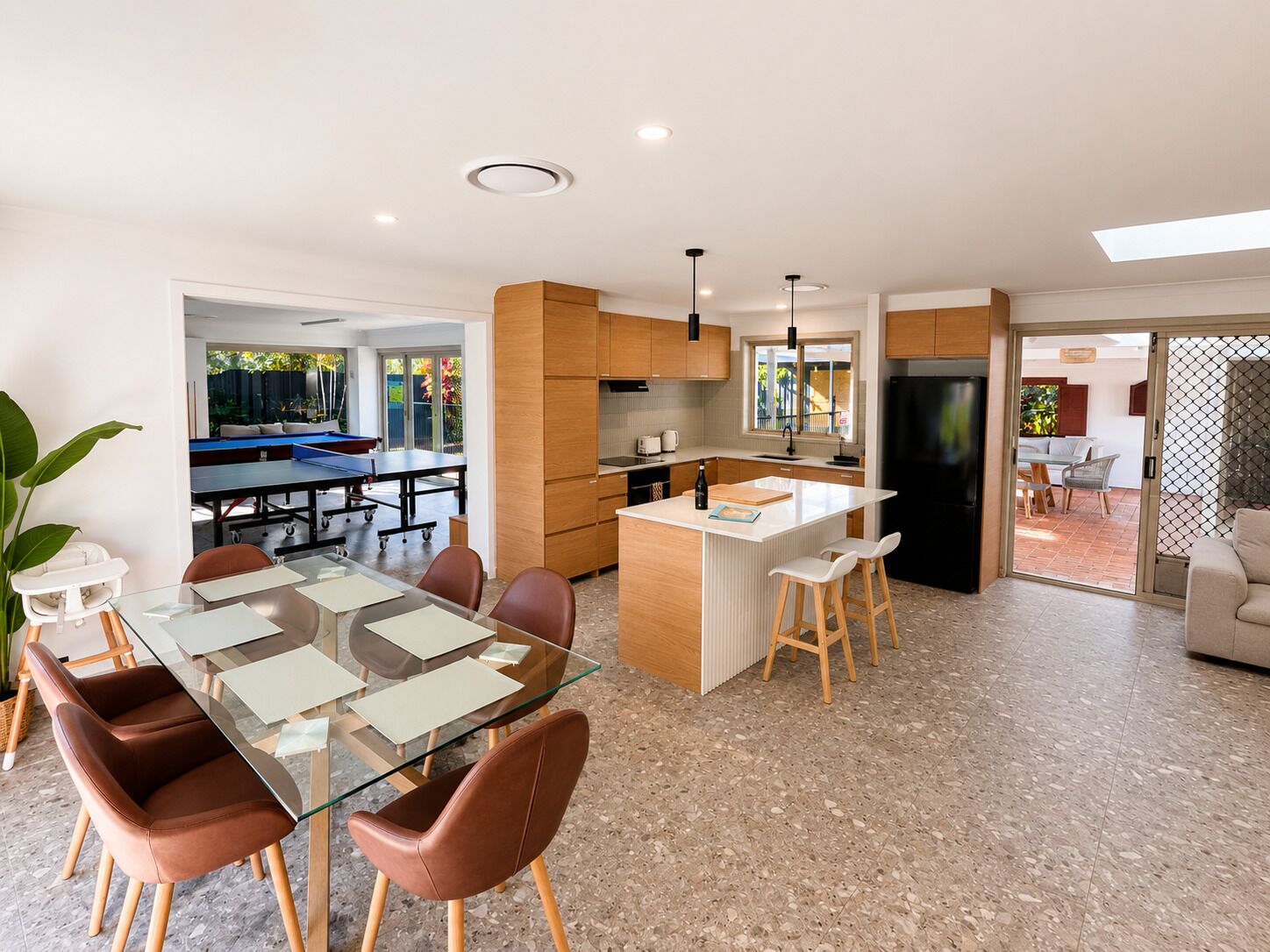 Une cuisine moderne dotée d’un îlot central, un coin repas avec une table et des chaises en verre, et une vue sur un espace de vie avec une piscine.