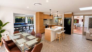 Une cuisine moderne dotée d’un îlot central, un coin repas avec une table et des chaises en verre, et une vue sur un espace de vie avec une piscine.