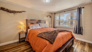 A bedroom with a wooden floor, a bed with an orange quilt, a nightstand with a lamp, and a window with curtains. - Beach Cabin #1, Private/Shared Beach - Cabin #1 (Lake Ozark)