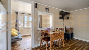 Uma sala de estar moderna com uma mesa de jantar de madeira, duas cadeiras de madeira, um sofá cinza com almofadas amarelas e uma TV fixada na parede.