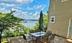 A patio with a table and chairs, overlooking a body of water with boats, trees, and a building with a door. - Mid-Century Cabin, Pet Friendly Mid-Century Cabin (Lake Ozark)