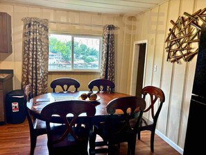 Uma sala de jantar com uma mesa redonda, seis cadeiras, uma janela com cortinas e um objeto decorativo na parede.