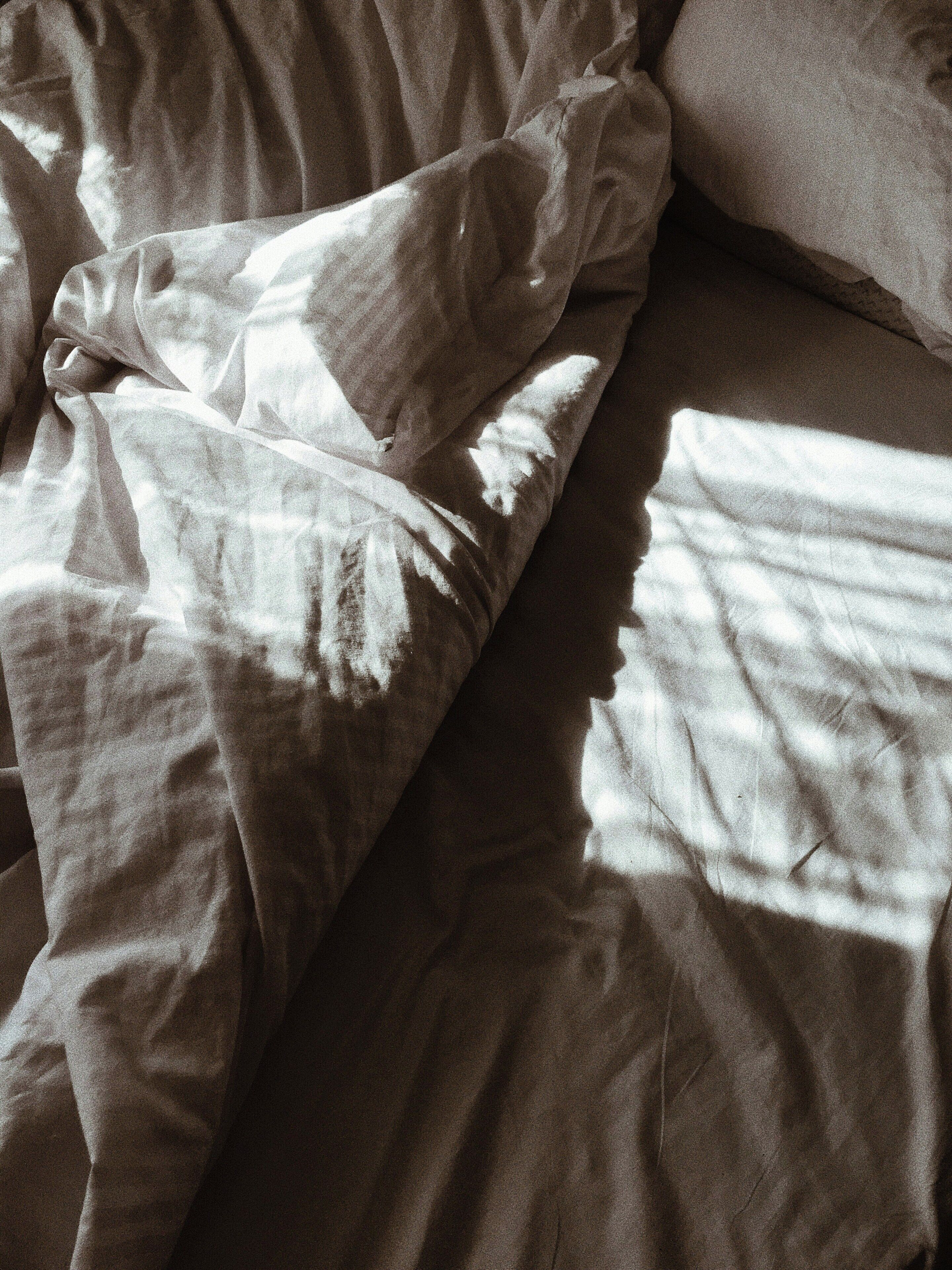 A bed with a white blanket and pillows, sunlight casting shadows on the bed.