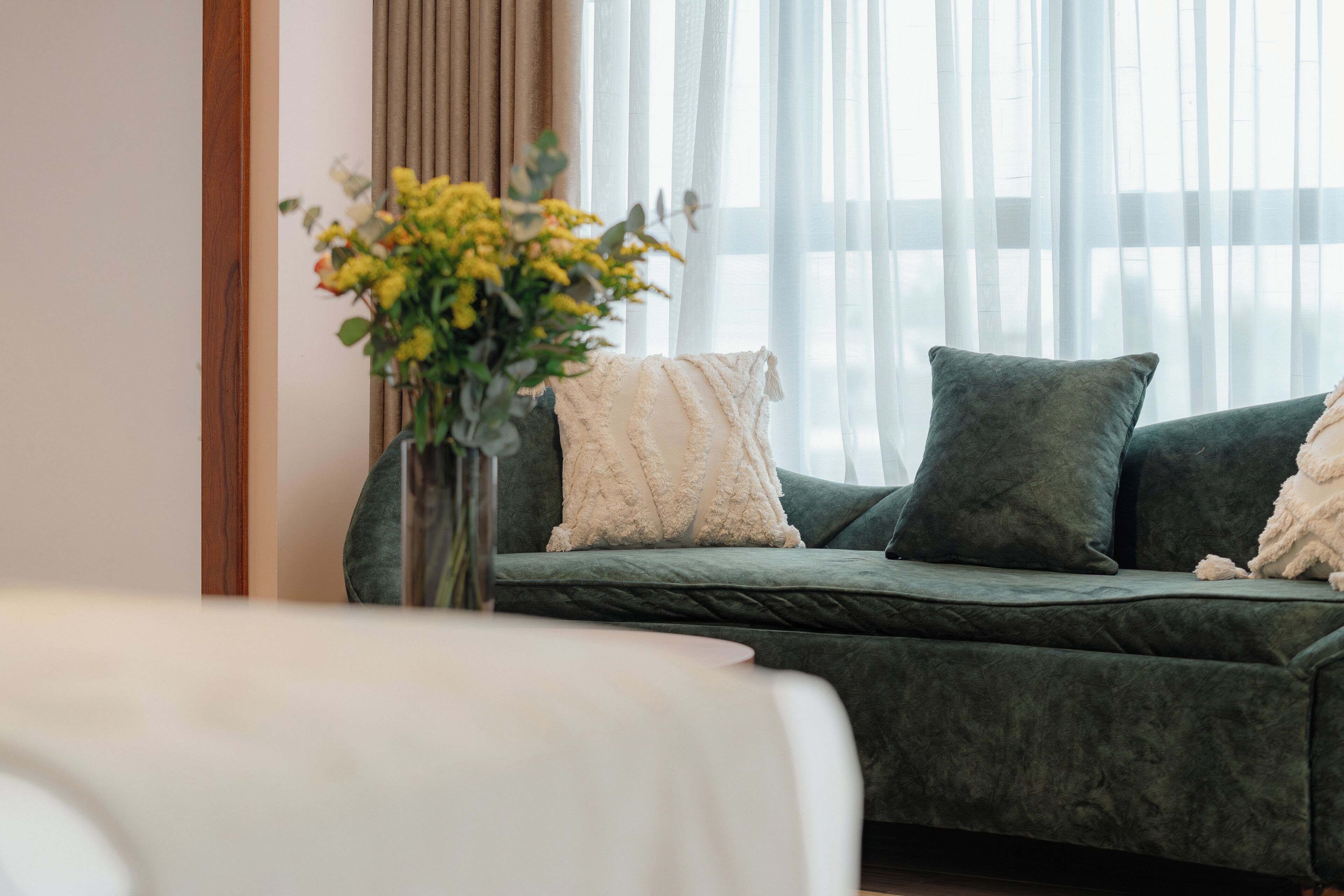 A green sofa with a vase of yellow flowers, a white cushion, and a window with sheer curtains.