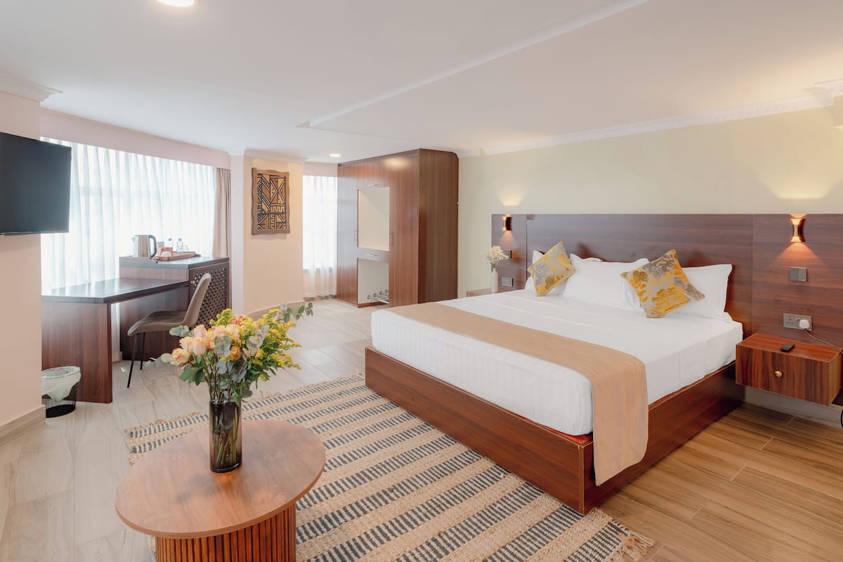Quarto de hotel com cama, mesa de cabeceira, televisão, escrivaninha, cadeira e um vaso com flores.