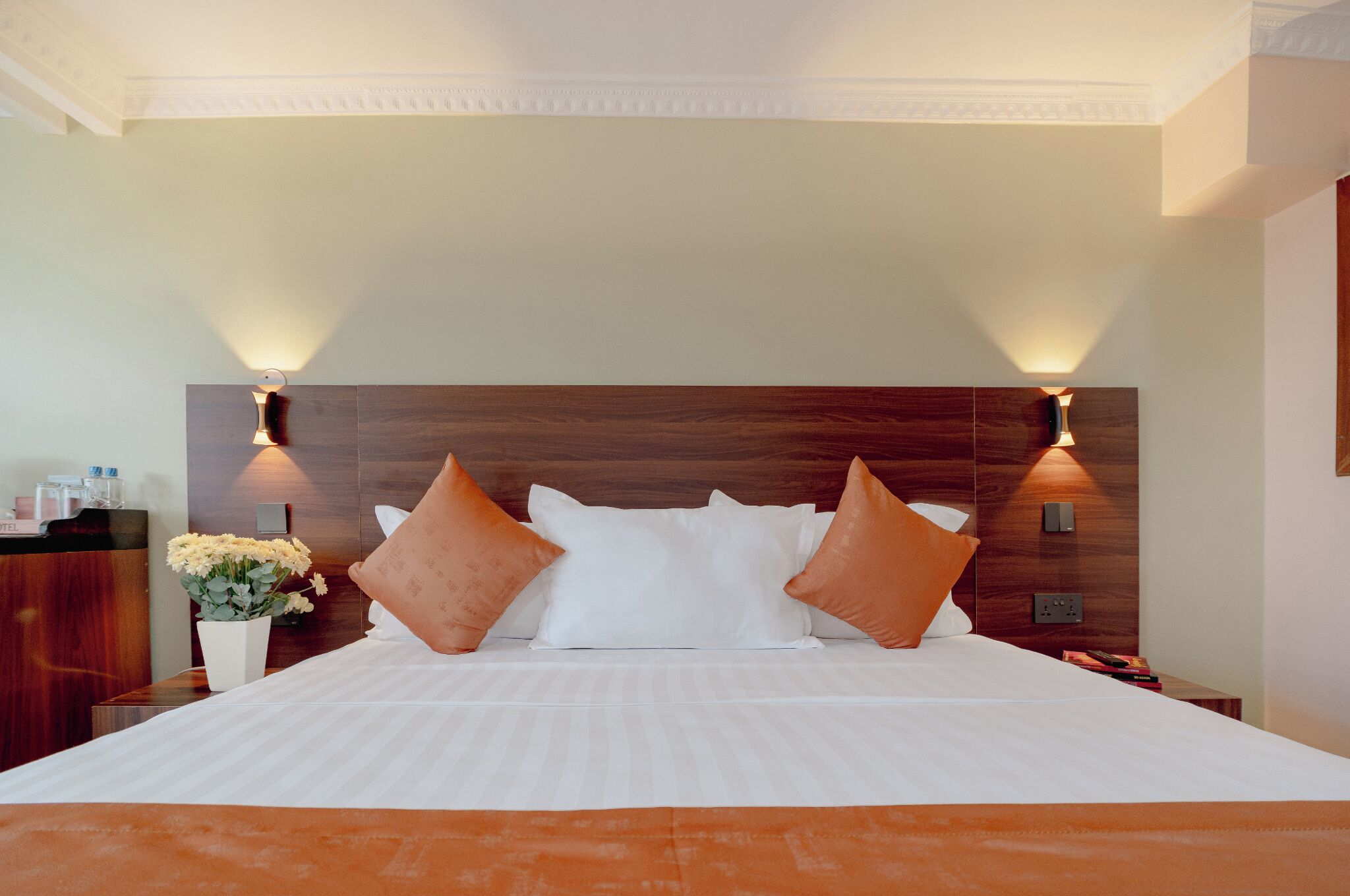 A hotel room with a large bed, two wall-mounted lamps, a wooden headboard, and a nightstand with a vase of flowers.
