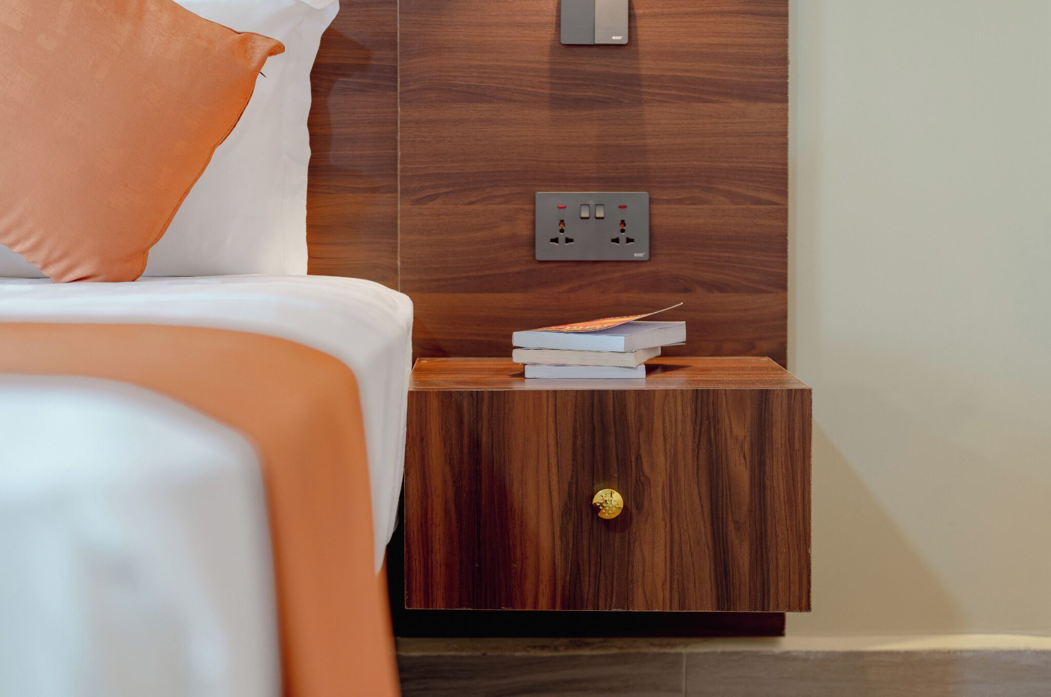 Un letto rifatto con un cuscino arancione, un comodino in legno con un libro e una presa elettrica, e un interruttore a muro.
