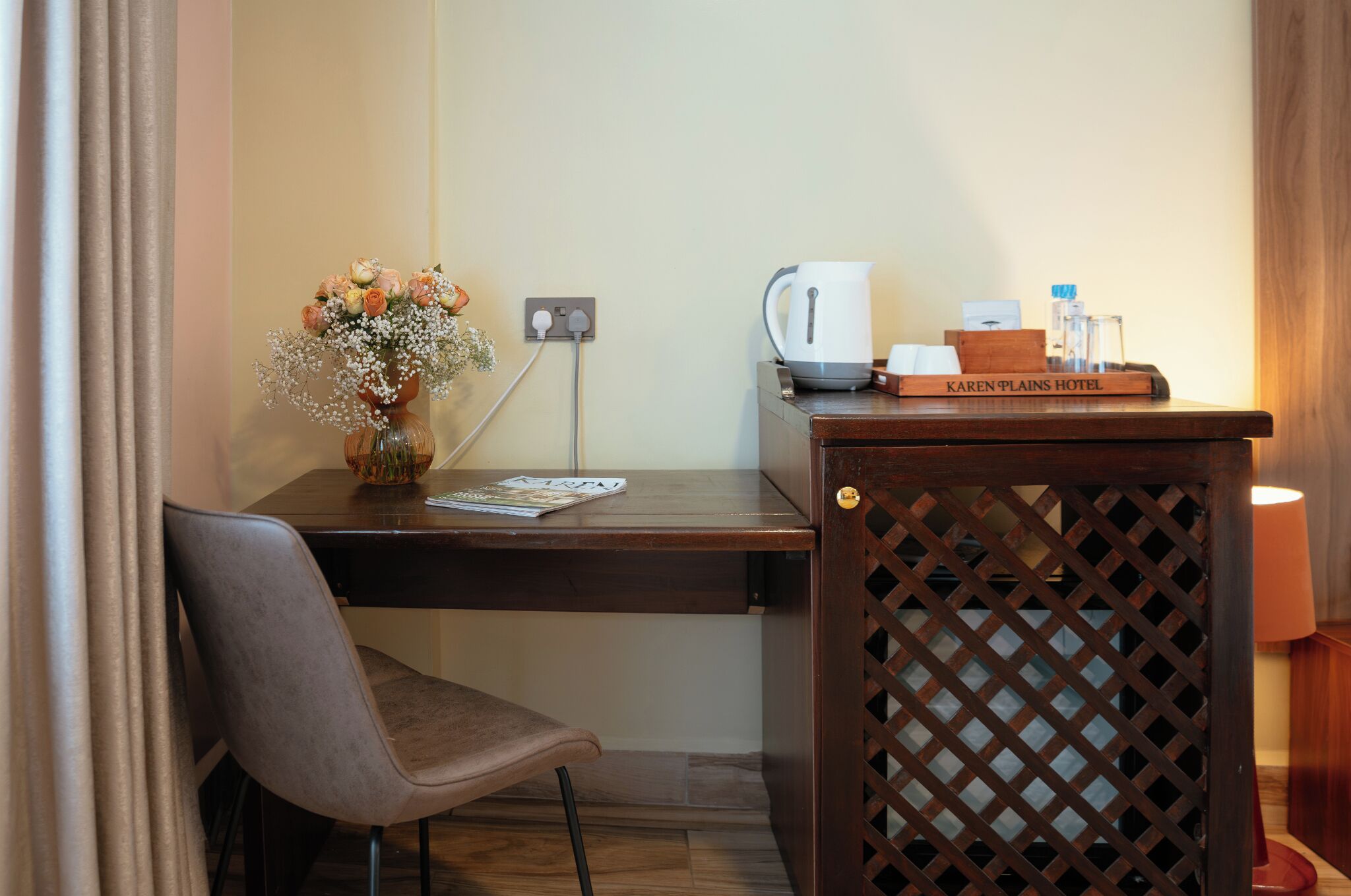 A hotel room with a wooden desk, a chair, a vase with flowers, a kettle, a tray with a book, and a lamp.