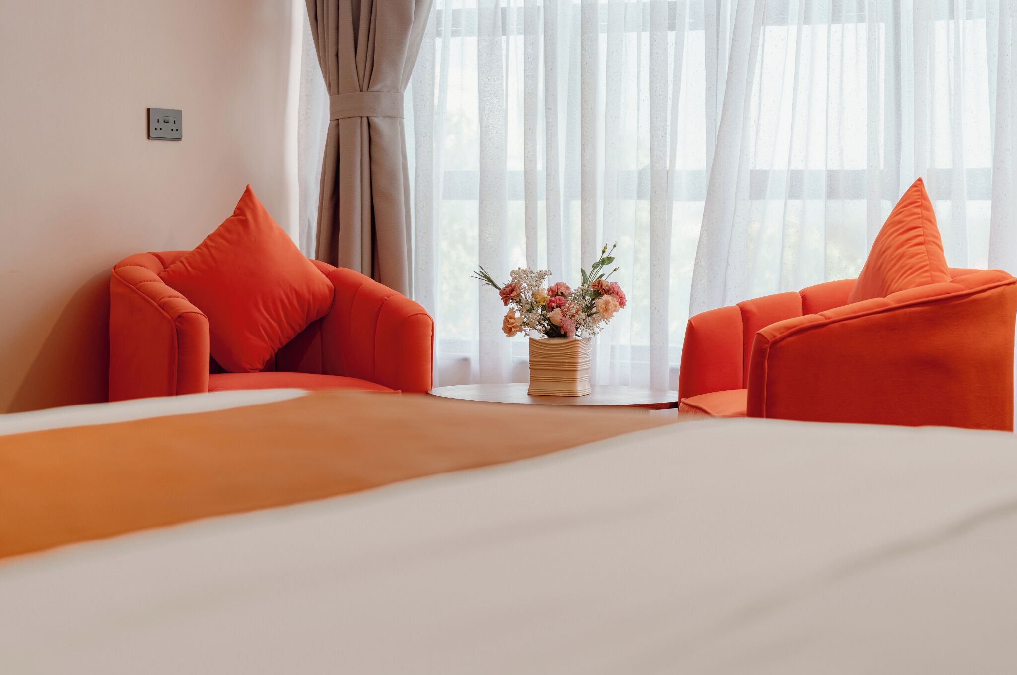 A hotel room with a bed, two orange chairs, a vase with flowers, and a window with sheer curtains.
