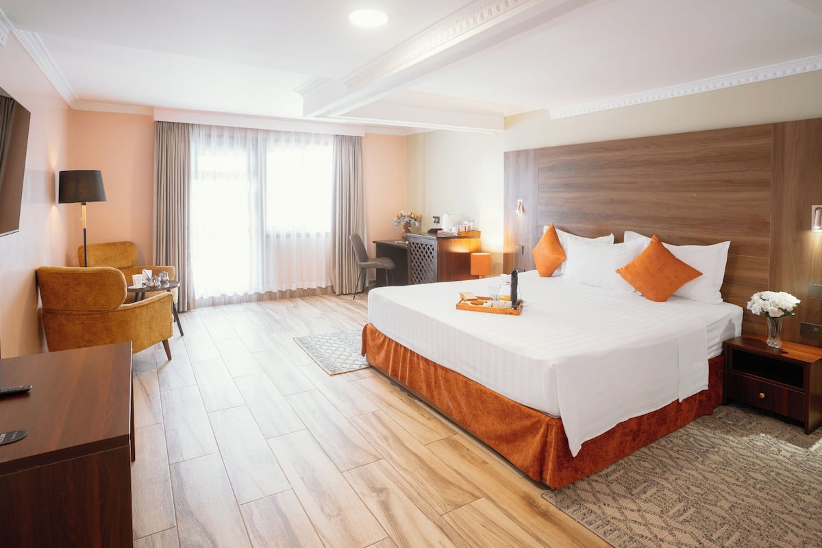 Quarto de hotel com uma cama grande, uma escrivaninha, uma cadeira, uma luminária de mesa, um criado-mudo e uma televisão.