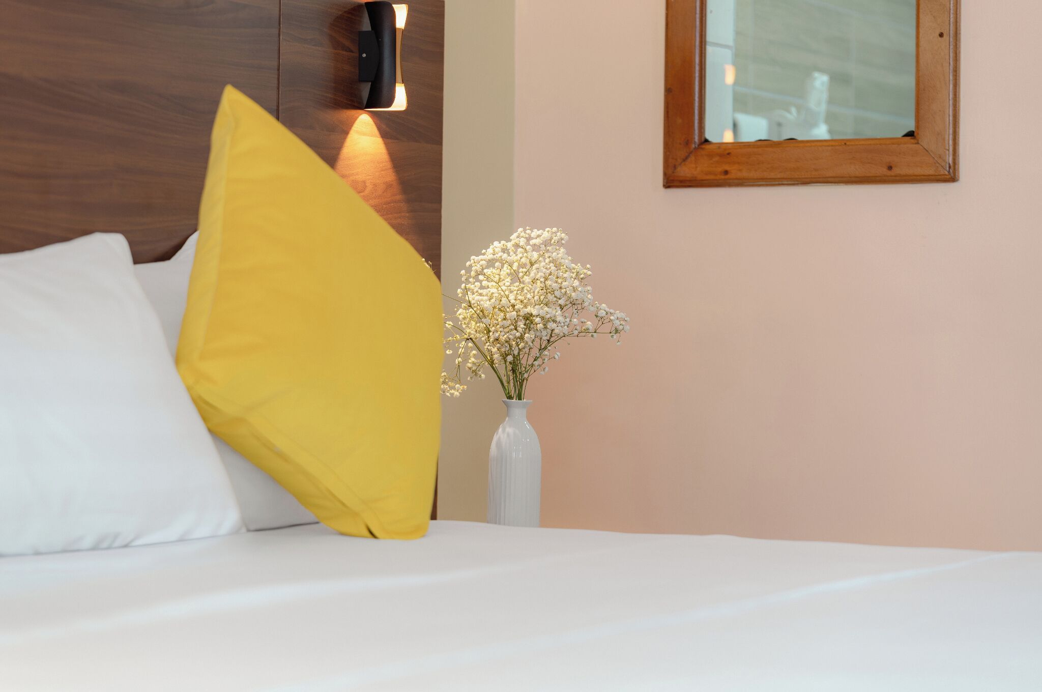 A bed with white and yellow pillows, a white vase with flowers, a wooden headboard, and a mirror on the wall.