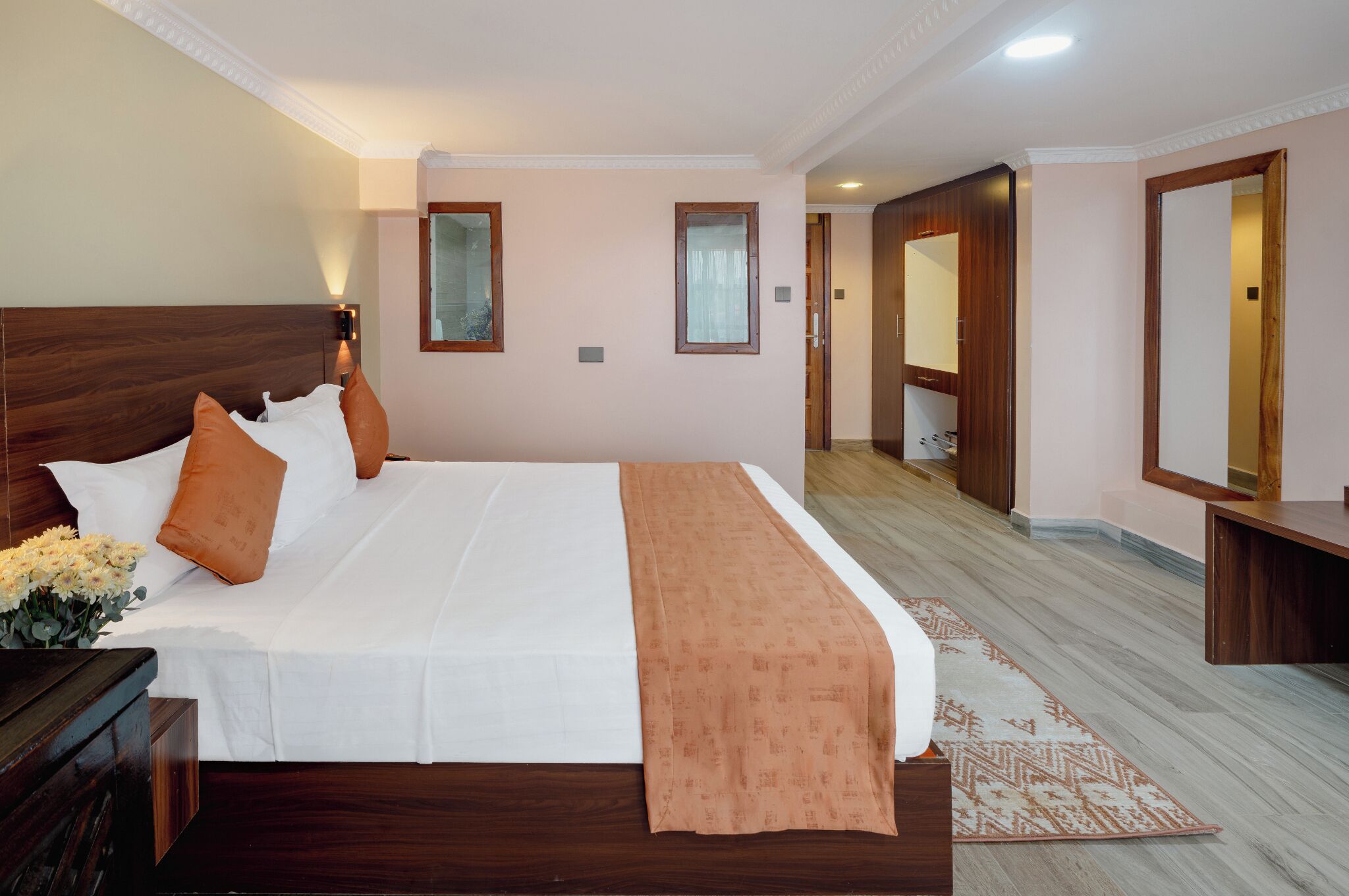A hotel room with a large bed, wooden headboard, a bedside table, a nightstand, a mirror, and a door leading to another room.