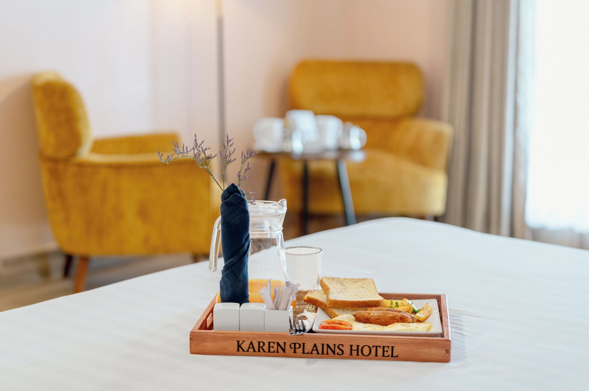 A hotel room with a bed, a tray with breakfast items, a pitcher, and a vase with flowers.
