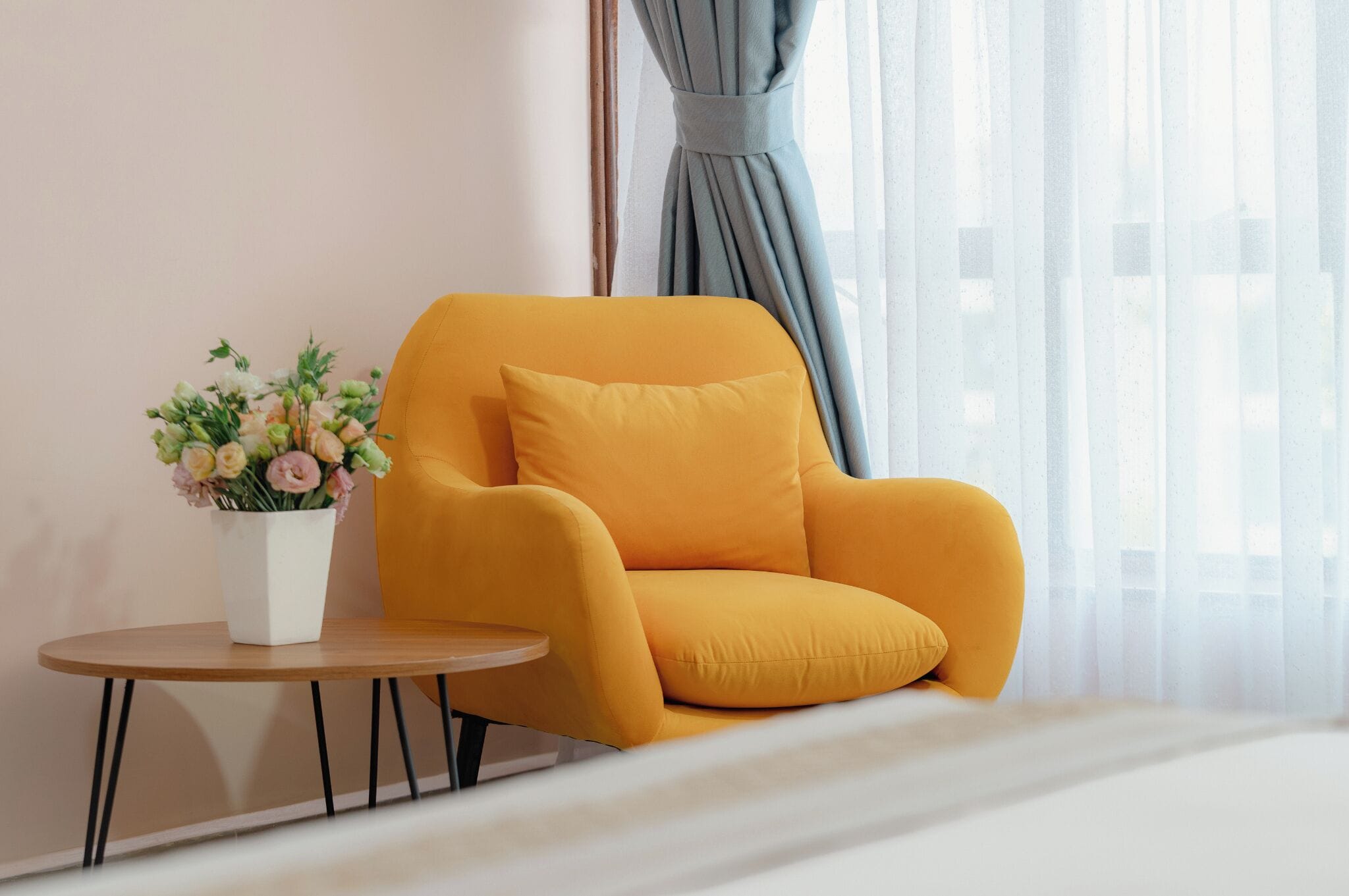 A room with a yellow armchair, a small round table, and a white vase with flowers.