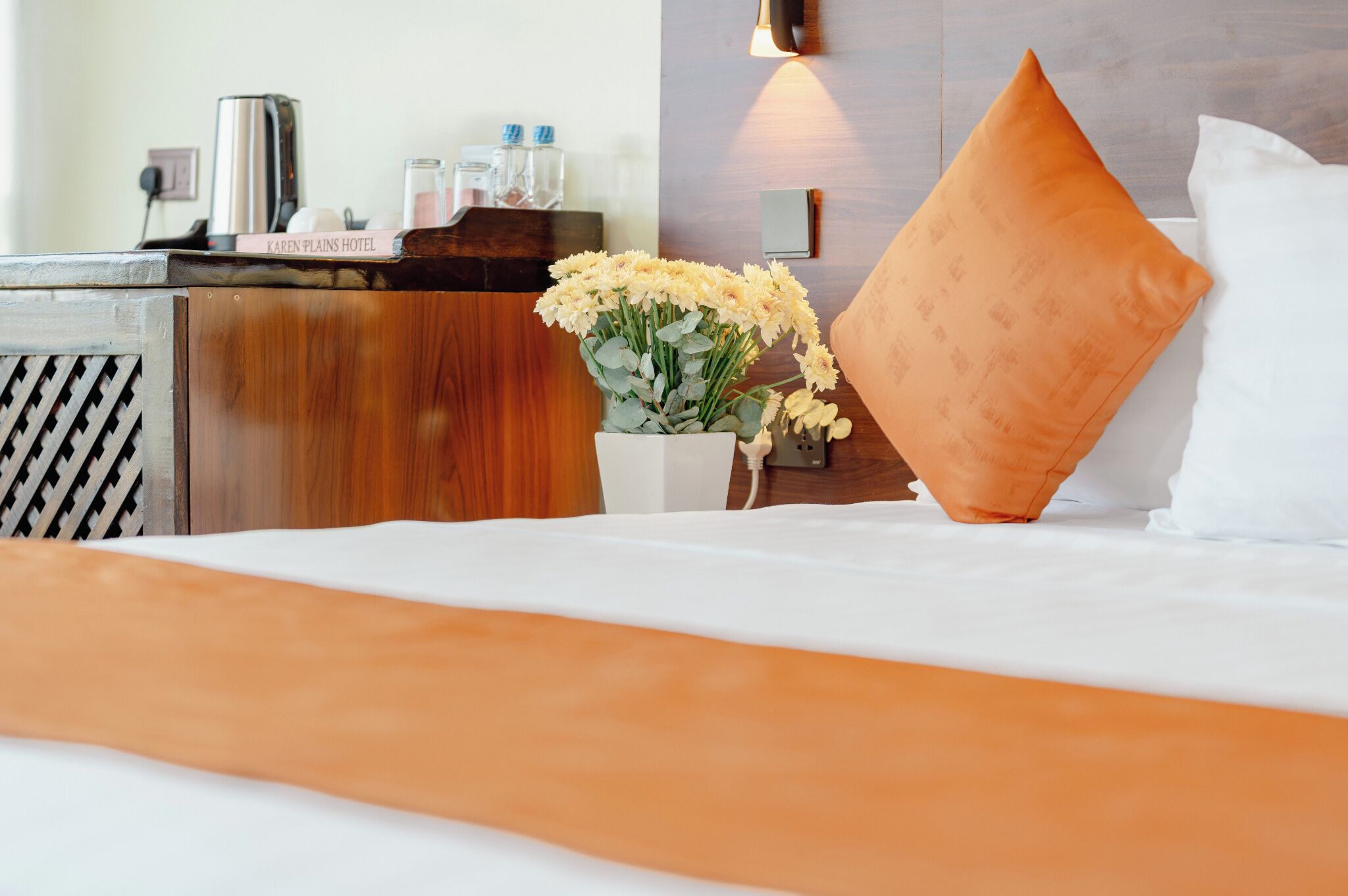 A hotel room with a bed, a bedside table with a vase of flowers, a lamp, and a sign reading 'Karen Plains Hotel'.