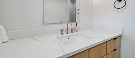 Un baño con lavamanos de mármol blanco, armarios de madera, un espejo grande y un aro para toallas en la pared.