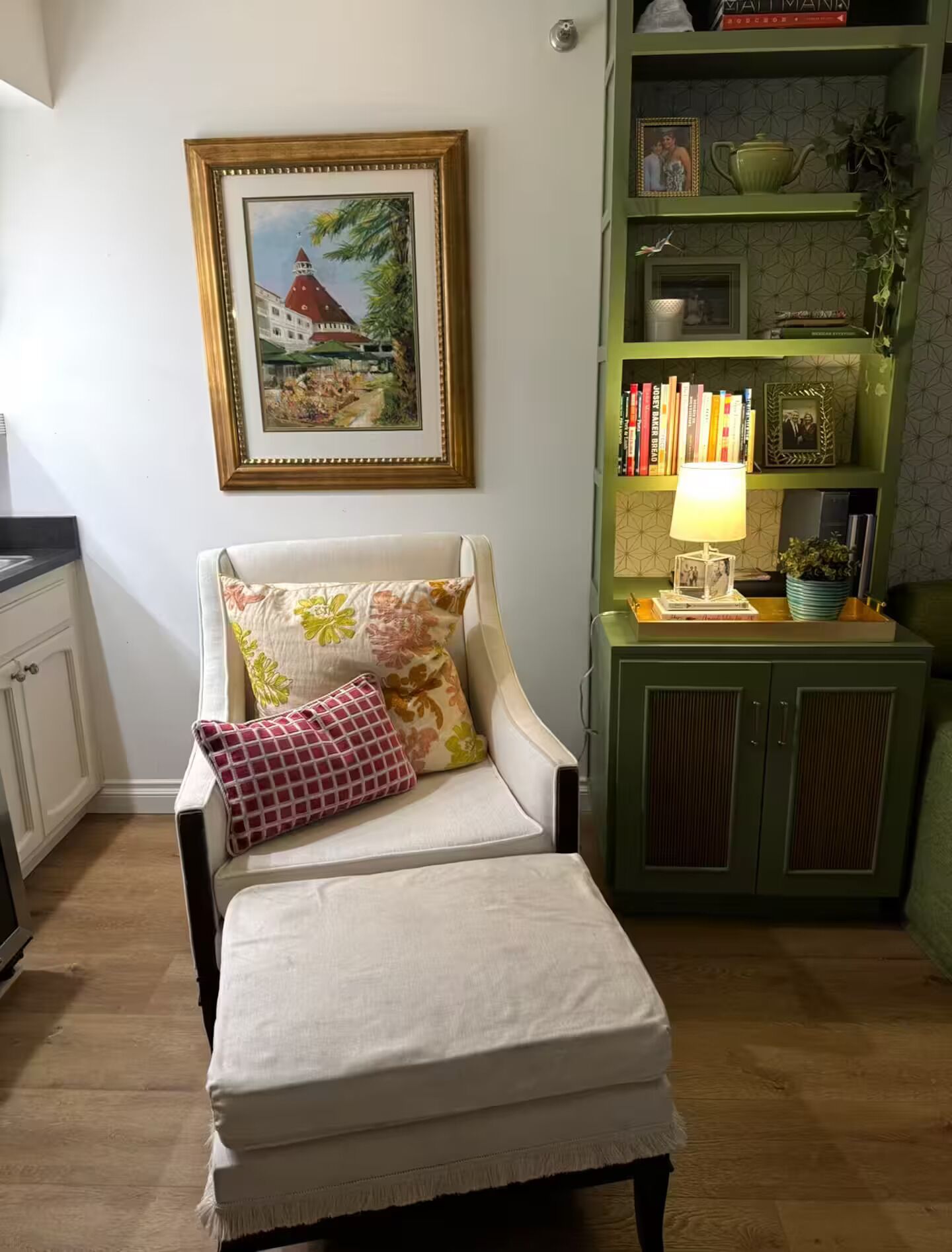 A cozy living room with a white armchair, a small ottoman, a bookshelf with books and decorative items, and a framed painting on the wall.