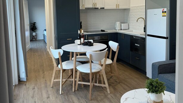 A modern kitchen with white cabinets, a built-in oven, and a refrigerator. There is a dining table with chairs and a small round table with a plant. - Kingdom Cottages (Gulgong)
