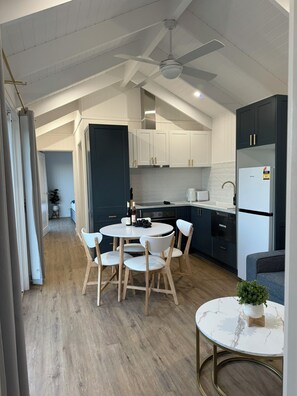 A modern kitchen with white cabinets, a built-in oven, and a refrigerator. There is a dining table with chairs and a small round table with a plant. - Kingdom Cottages (Gulgong)