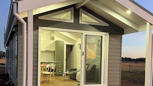 A modern, single-story house with a wooden deck and a sliding glass door leading to an interior with a kitchen and living area. - Kingdom Cottages (Gulgong)