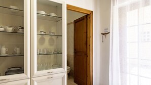 A room with a glass-fronted cabinet containing dishes and glasses, a wooden door, and a window with sheer curtains.