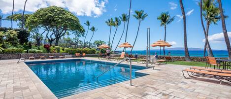 A pool area with lounge chairs and umbrellas, surrounded by palm trees and a view of the ocean.