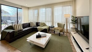 Un salon moderne avec un canapé d’angle, une table basse, un téléviseur et une grande fenêtre agrémentée de rideaux.
