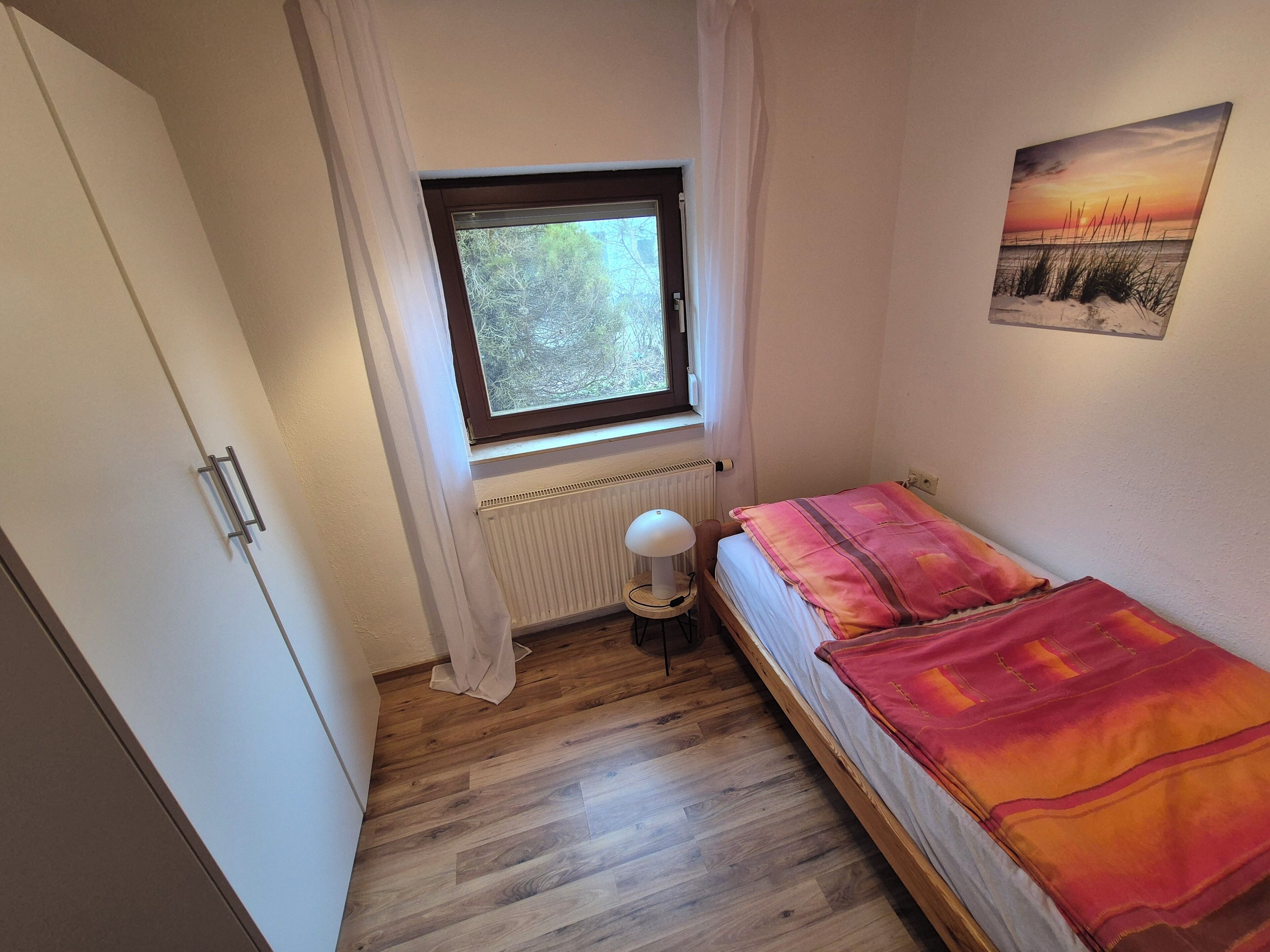 A small bedroom with a single bed, a window with curtains, a radiator, and a lamp.
