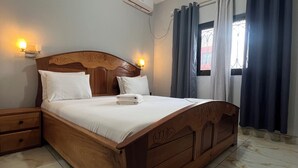 Een netjes opgemaakt bed met witte lakens, een houten hoofdbord en een nachtkastje met lades.