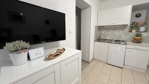 Una cocina moderna con gabinetes blancos, horno empotrado y lavamanos. Un televisor de pantalla plana está montado sobre la encimera, y hay una pequeña planta en maceta sobre la misma.