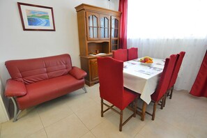 A dining area with a red sofa, wooden cabinet, and a table set with chairs and a fruit bowl. - Apartmani Vela Makarska (Makarska)