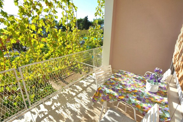 A balcony with a table and chairs, a white railing, and a view of greenery and parked cars. - Apartmani Vela Makarska (Makarska)