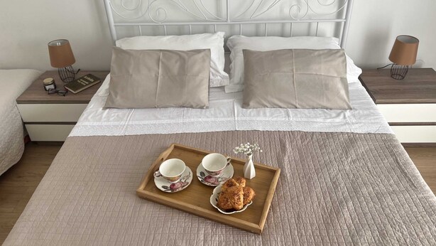 Una cama bien hecha con cabecera blanca, dos mesitas de noche con lámparas y una bandeja con tazas y bollería sobre la cama.