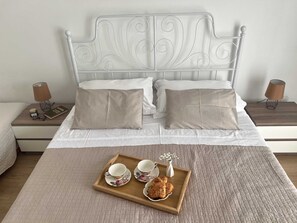 A neatly made bed with a white headboard, two bedside tables with lamps, and a tray with cups and pastries on the bed. - Apartmani Vela Makarska (Makarska)