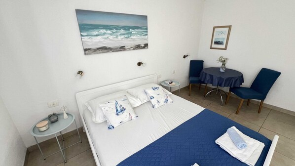 A hotel room with a bed, a small table with a vase, a chair, a round table with a chair, and a painting on the wall. - Apartmani Vela Makarska (Makarska)
