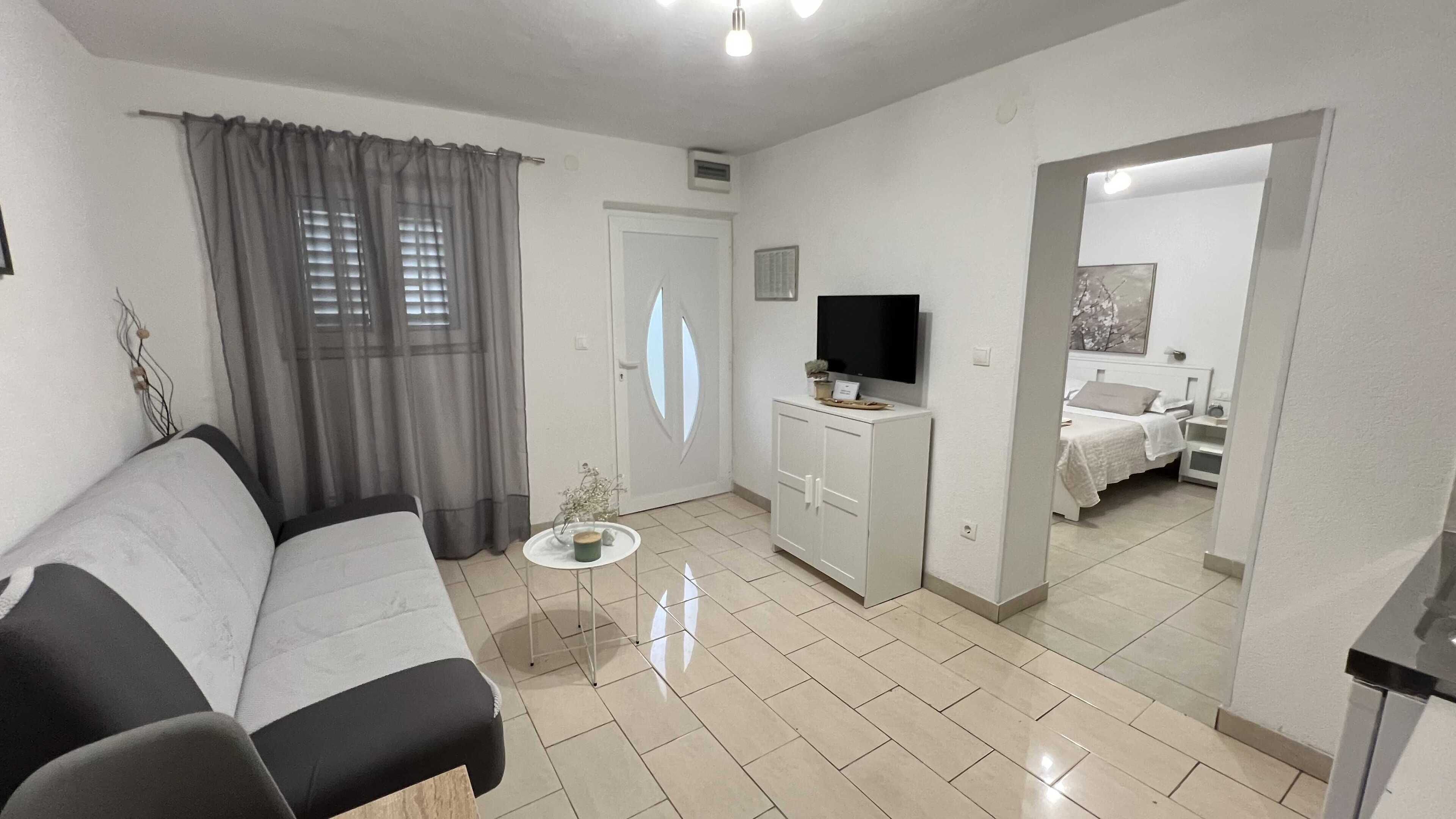 A modern living room with a sofa, a small table with a plant, a TV on a cabinet, and a bedroom visible through an open door.