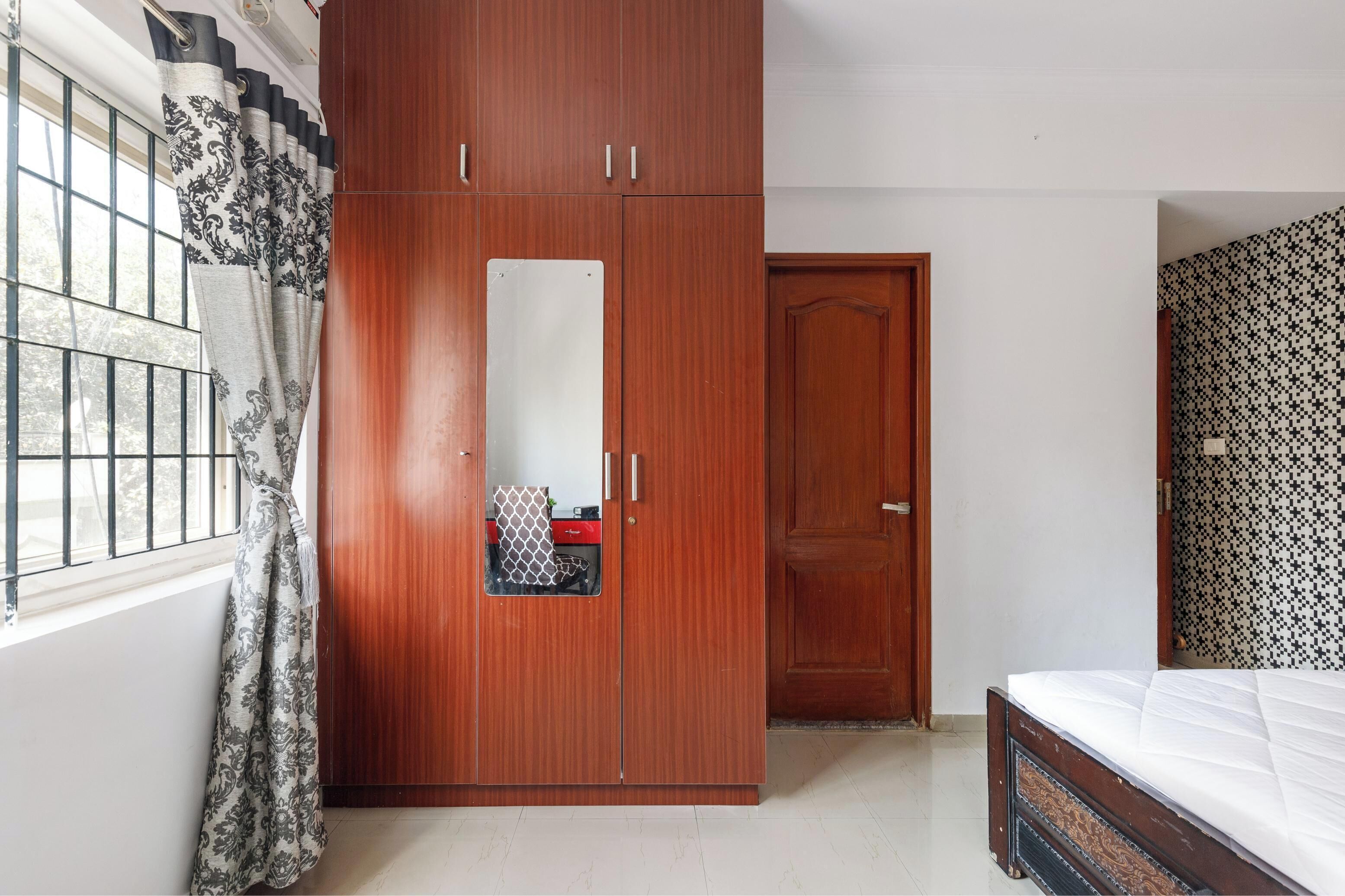 A bedroom with a wooden wardrobe, a bed, and a window with patterned curtains.